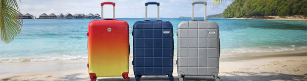 Three suitcases in red, blue, and gray on a sandy beach with palm trees and ocean in the background.